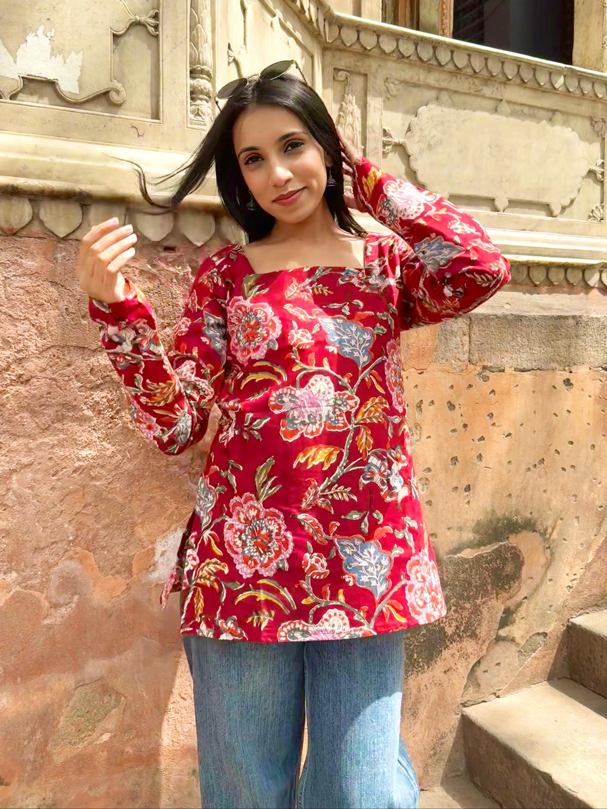 Woman wearing a red floral blouse standing in front of an old architectural wall.