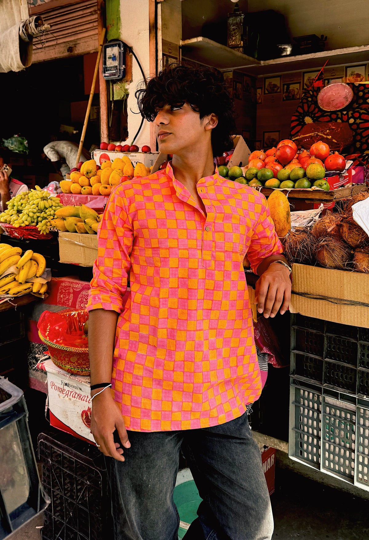 Person wearing a colorful checkered shirt in a market setting with fruits and vegetables.
