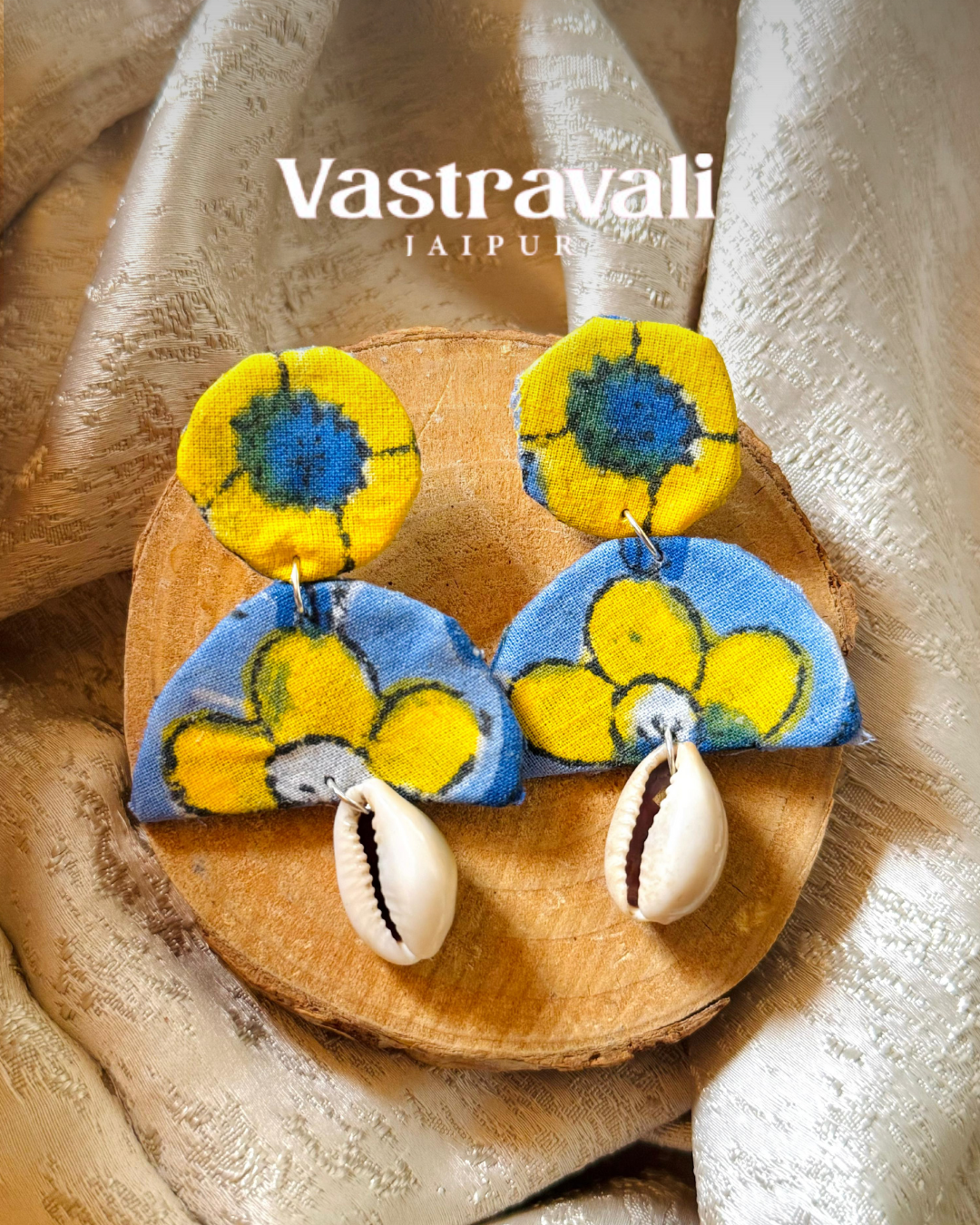 Pair of colorful earrings with cowrie shells on a wooden board, branded 'Vastravali'.