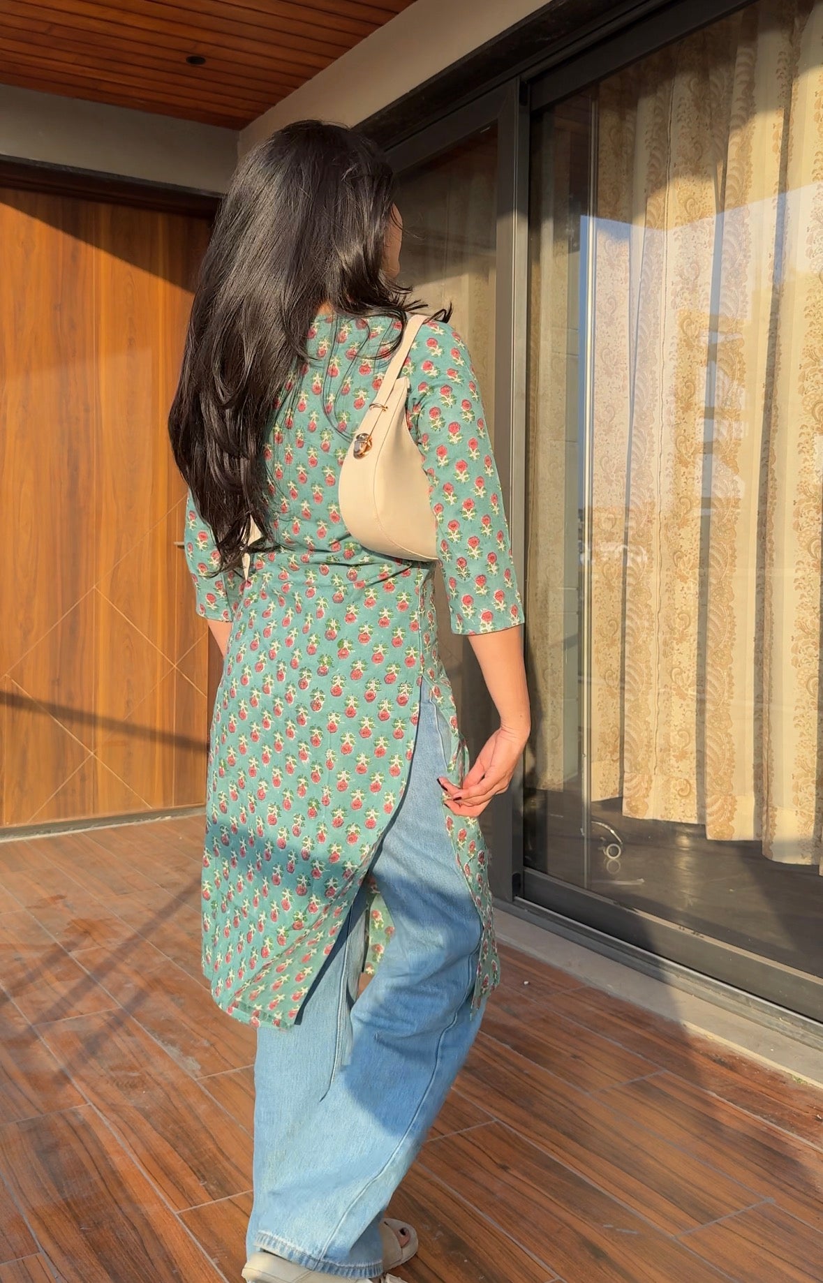 Woman in a green patterned shirt and blue jeans walking on a wooden floor.