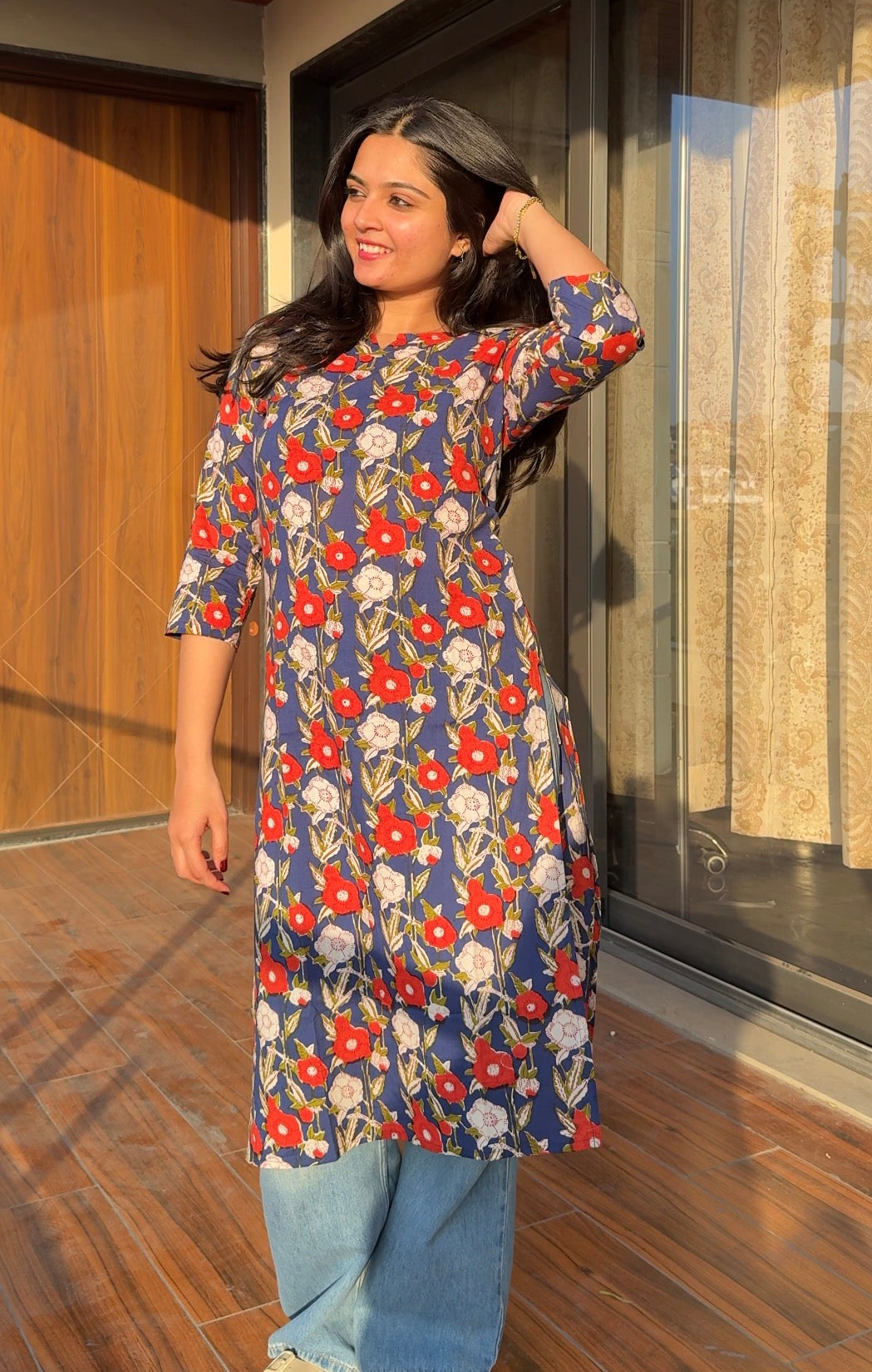 Woman wearing a floral dress standing indoors with wooden flooring and a glass door in the background.
