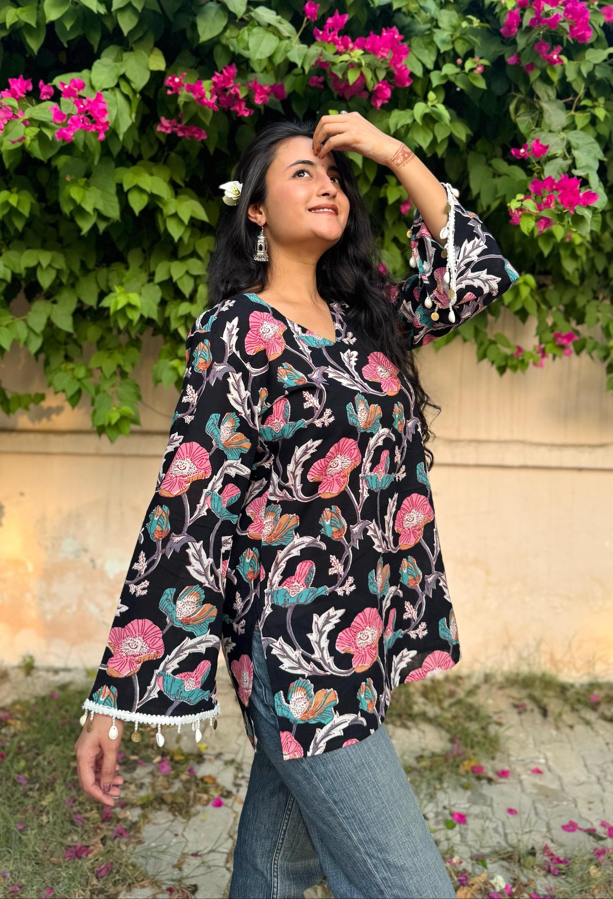 A woman standing near a wall with floral surroundings, wearing a black short kurti with floral hand-block printing and flared sleeves.