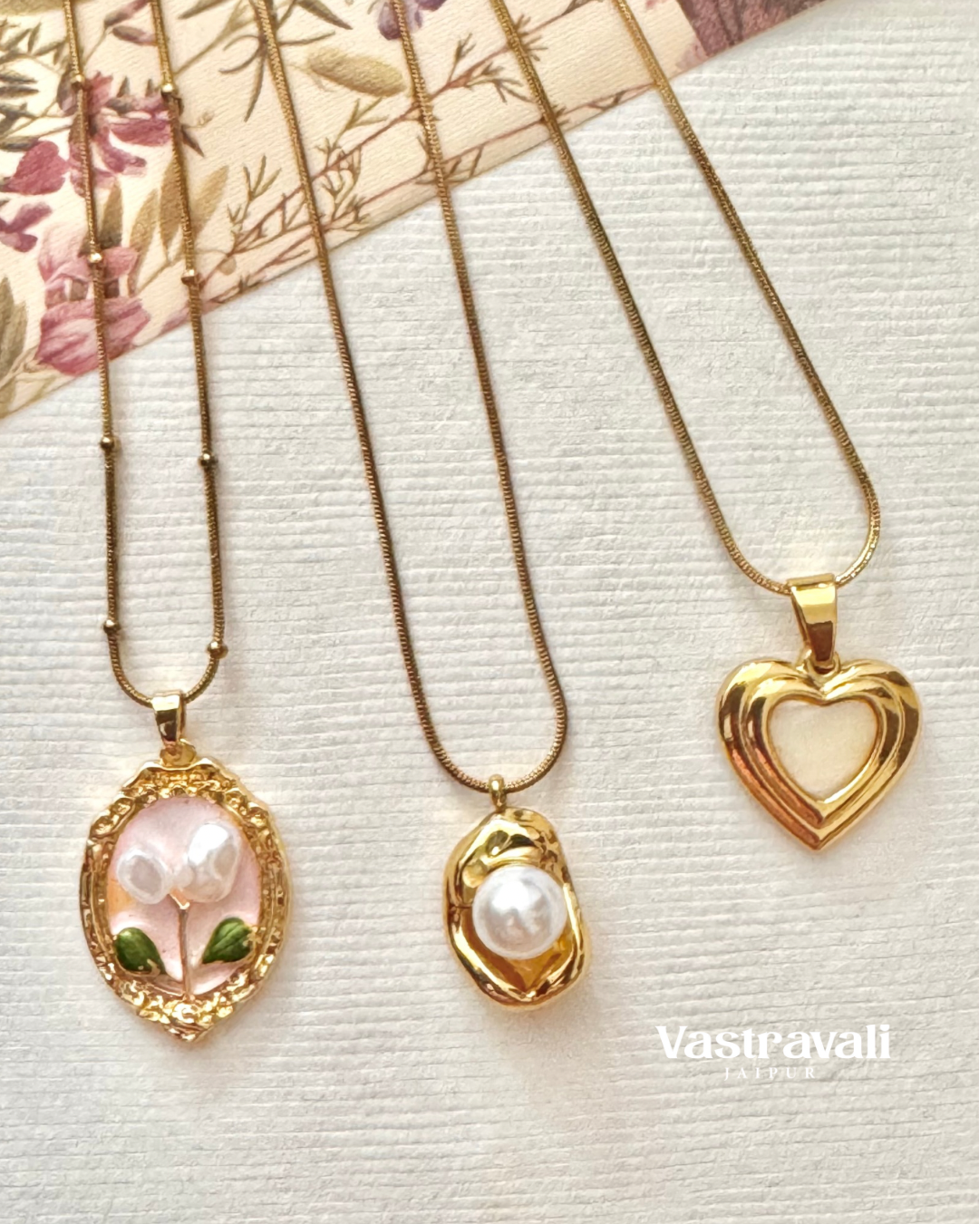 Three gold necklaces with pendants on a textured surface, featuring the brand 'Vasavalli'.