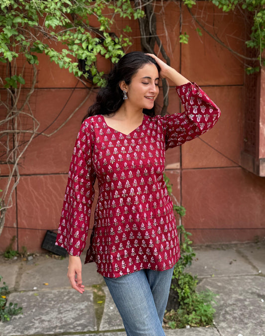 A woman wearing a red, patterned top kurti and blue jeans stands outdoors, surrounded by greenery and a brown wall in the background.