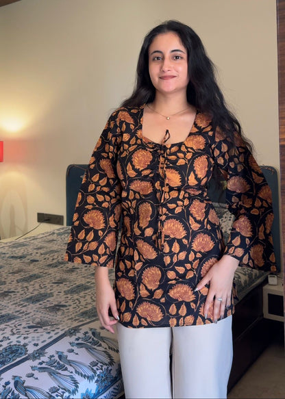Woman wearing a black and orange patterned top in a room with a bed and lamp.