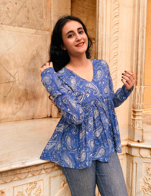 A woman standing with one hand on a railing, wearing a blue paisley hand-block printed cotton top with long sleeves.