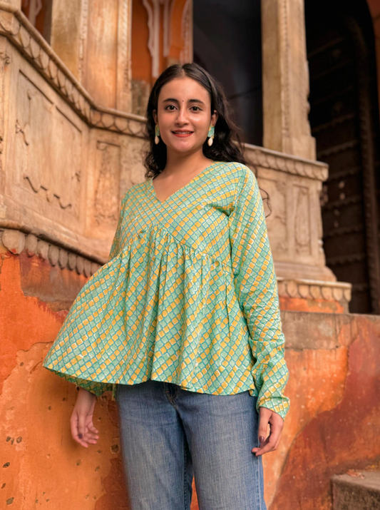 A woman wearing a green and yellow patterned top with a round neckline, standing in front of a colorful wall.