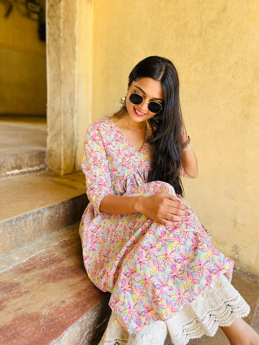 A woman sitting on stairs wearing a floral pink long kurti with white and pink flowers, paired with sunglasses and white trousers.