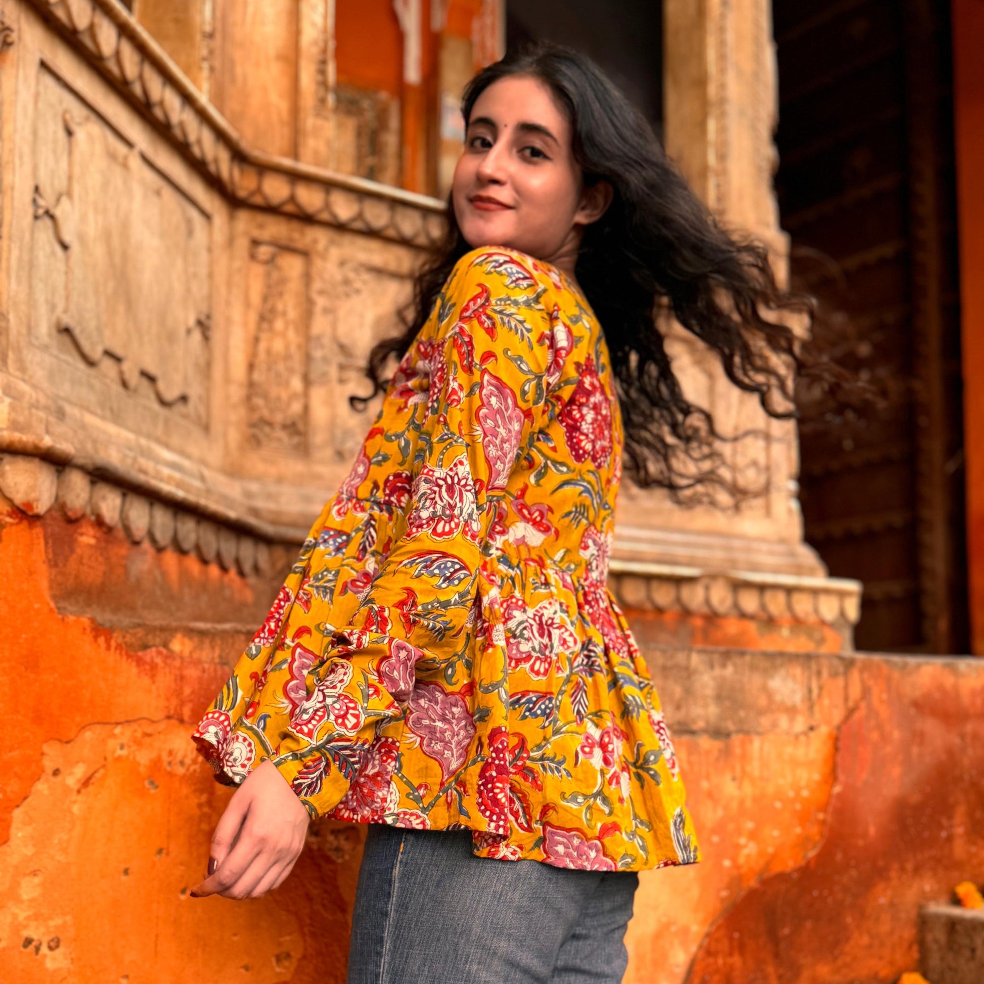 A person wearing a mustard yellow floral top with a flared cut, standing in front of a colorful wall.