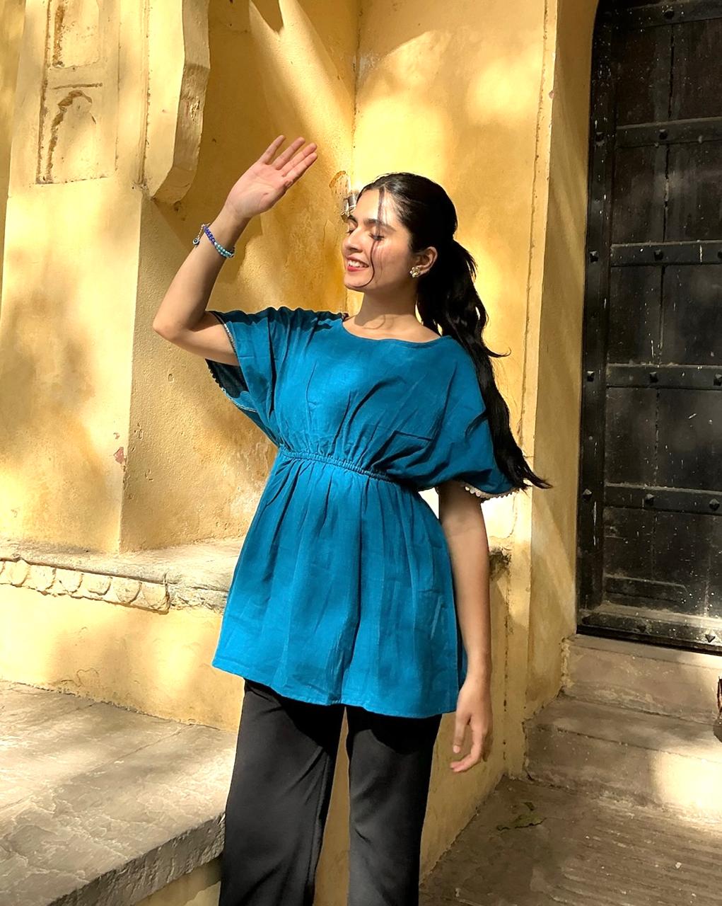 Woman in a blue top and black pants waving in front of a yellow wall with a large black door.