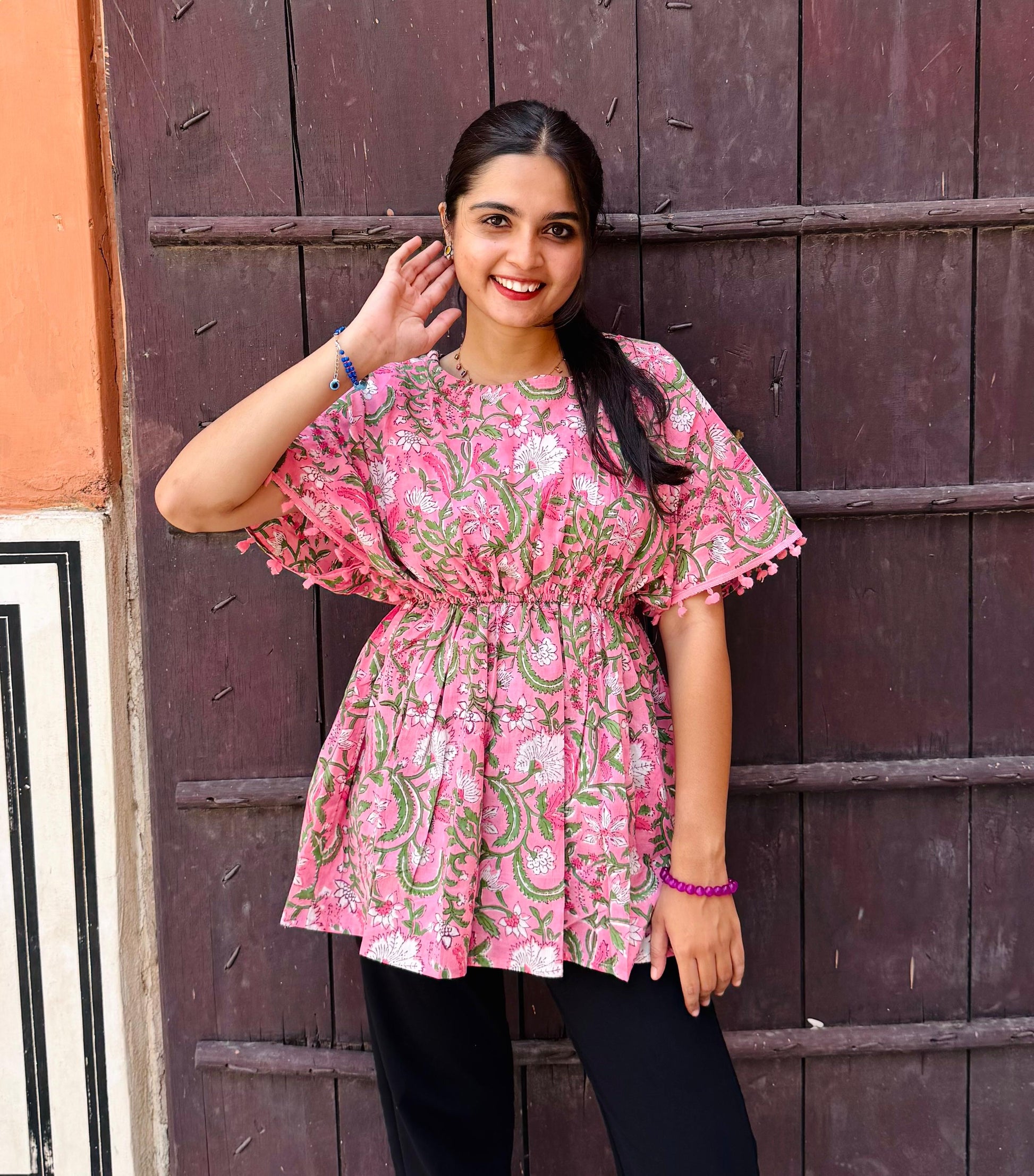 A woman standing against a wooden backdrop wearing a pink floral print top with short sleeves and a round neckline.