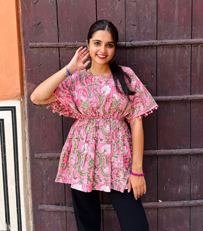 A woman standing against a wooden backdrop wearing a pink floral print top with short sleeves and a round neckline.