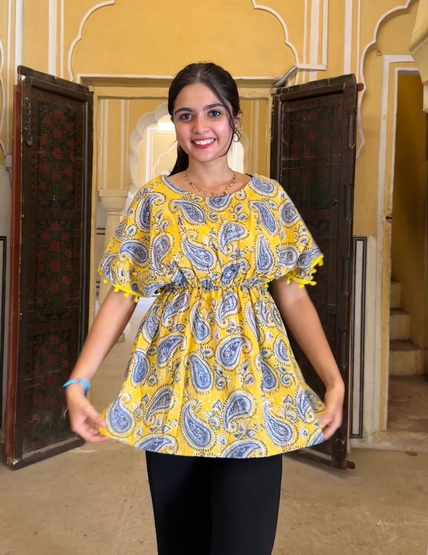 A woman standing in an indoor setting wearing a yellow top with a paisley floral pattern, paired with black pants.