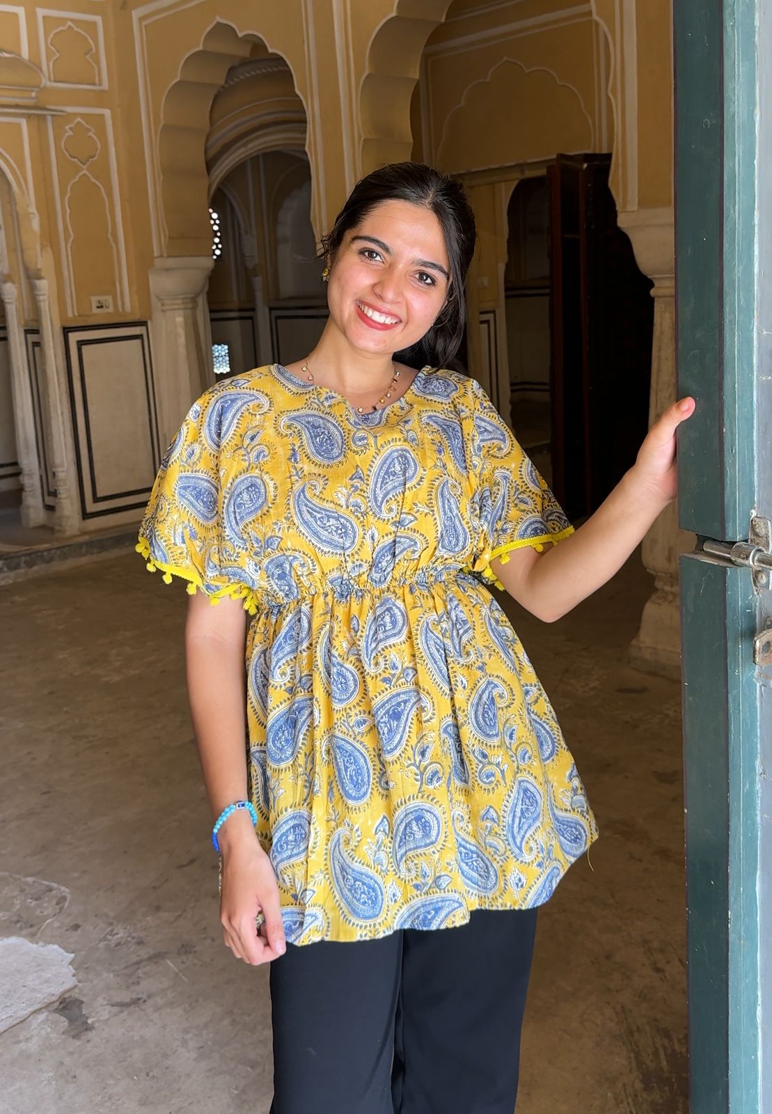 Woman wearing a yellow and blue patterned blouse standing in an ornate indoor setting.