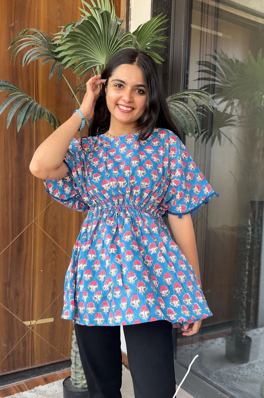 A woman smiling and posing in a blue floral print top with short sleeves, paired with black pants.