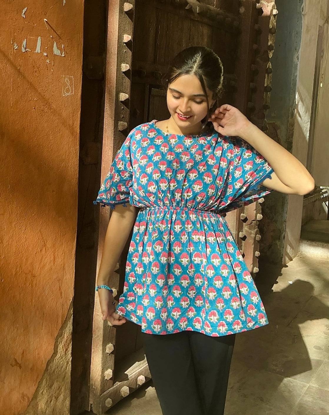 Woman wearing a blue patterned top and black pants standing in front of a wooden door.