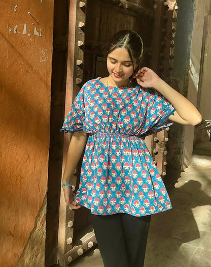 Woman wearing a blue patterned top and black pants standing in front of a wooden door.
