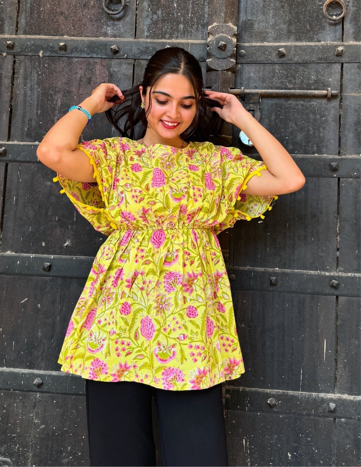 A woman standing against a dark background, wearing a yellow floral print top with short sleeves and a round neckline.