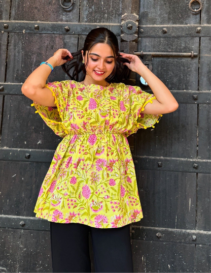 A woman standing against a dark background, wearing a yellow floral print top with short sleeves and a round neckline.