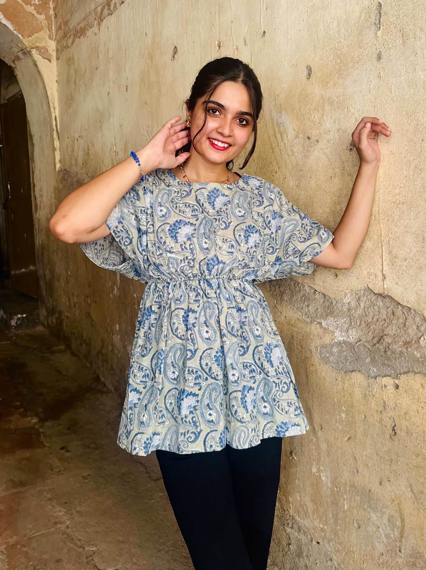 A woman posing against a textured wall, wearing a blue floral print top with short sleeves and a round neckline.