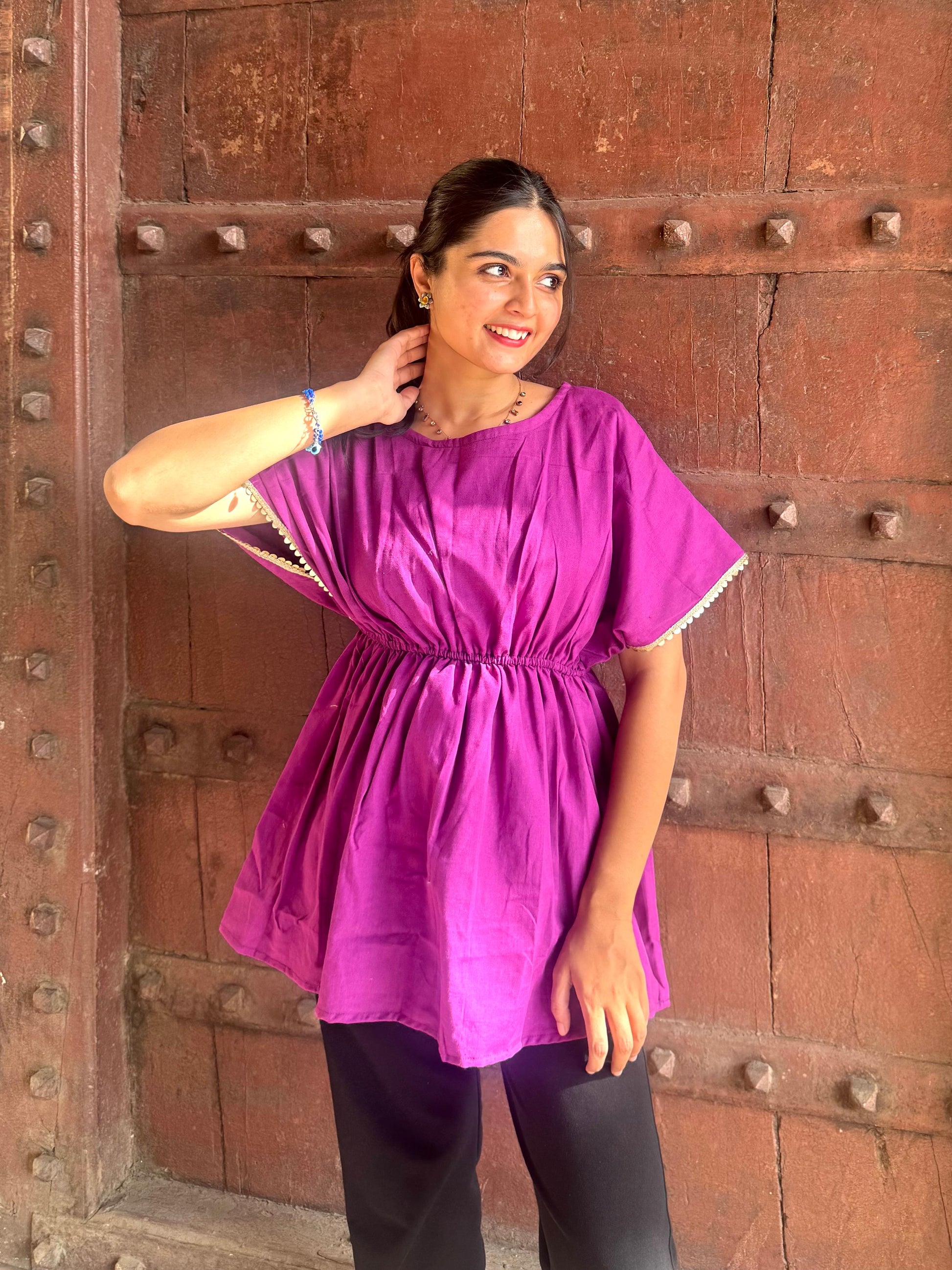 A person wearing a purple tunic top with short sleeves, standing in front of a dark background.