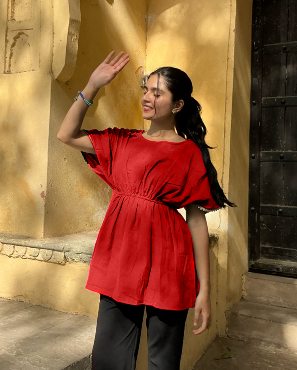 Woman in a red top and black pants standing against a yellow wall with architectural elements.