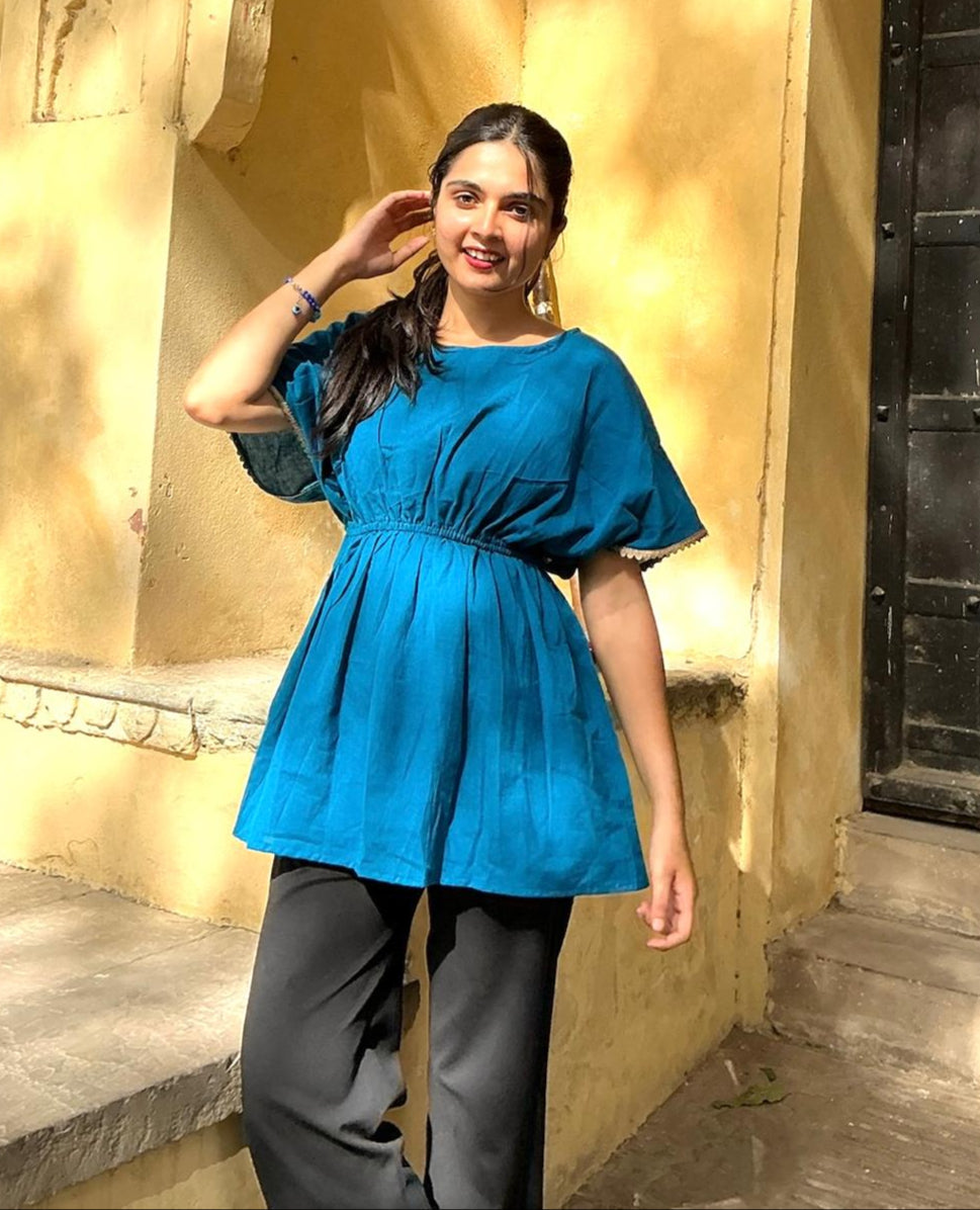 A woman wearing an ocean blue short-sleeved top with a round neckline and pleated detail at the waist, paired with dark pants.