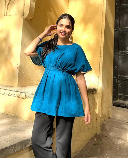 A woman wearing an ocean blue short-sleeved top with a round neckline and pleated detail at the waist, paired with dark pants.