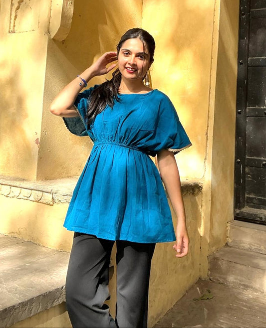 A woman wearing an ocean blue short-sleeved top with a round neckline and pleated detail at the waist, paired with dark pants.