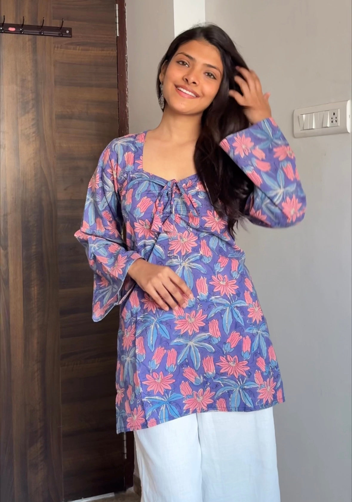 Woman wearing a blue floral top in a room with wooden door and light-colored wall.