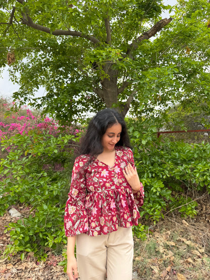 Woman in a red floral blouse standing outdoors with greenery and pink flowers in the background