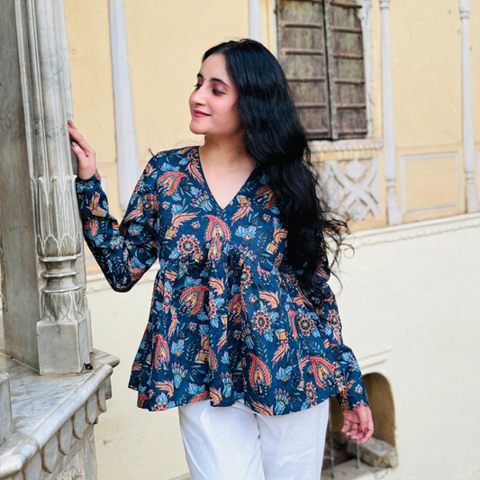 A woman standing near a building column, wearing a dark blue paisley print top with long sleeves and a V-neck neckline.