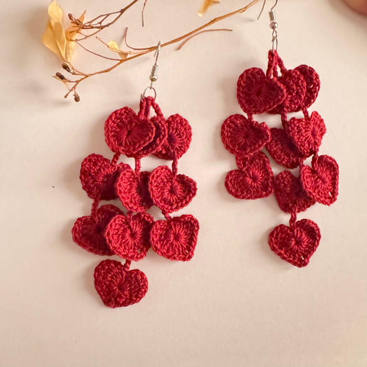 A pair of red, heart-shaped crochet earrings displayed against a white background with a few dried leaves for decoration.