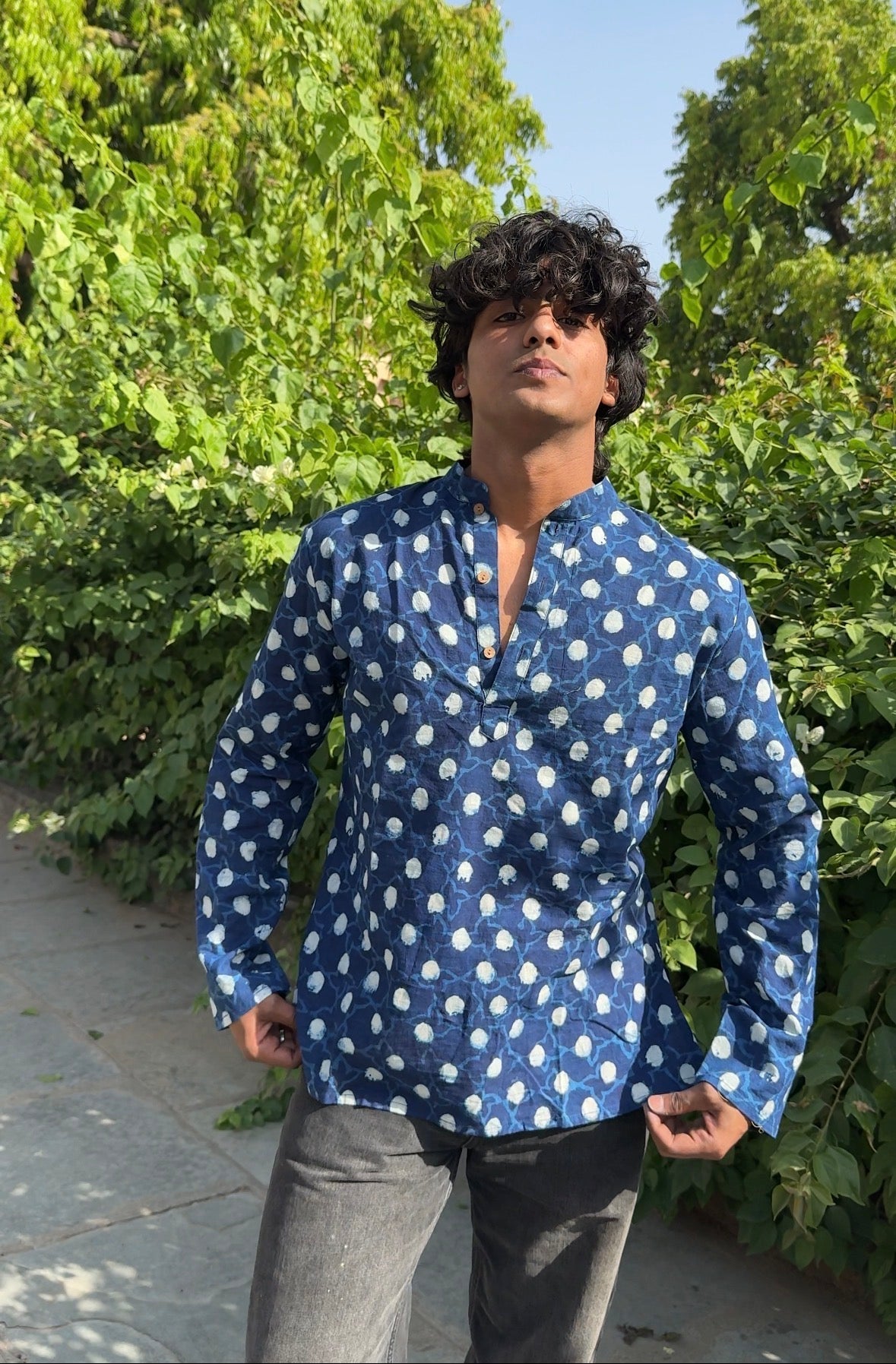Person wearing a blue patterned shirt standing outdoors with greenery in the background