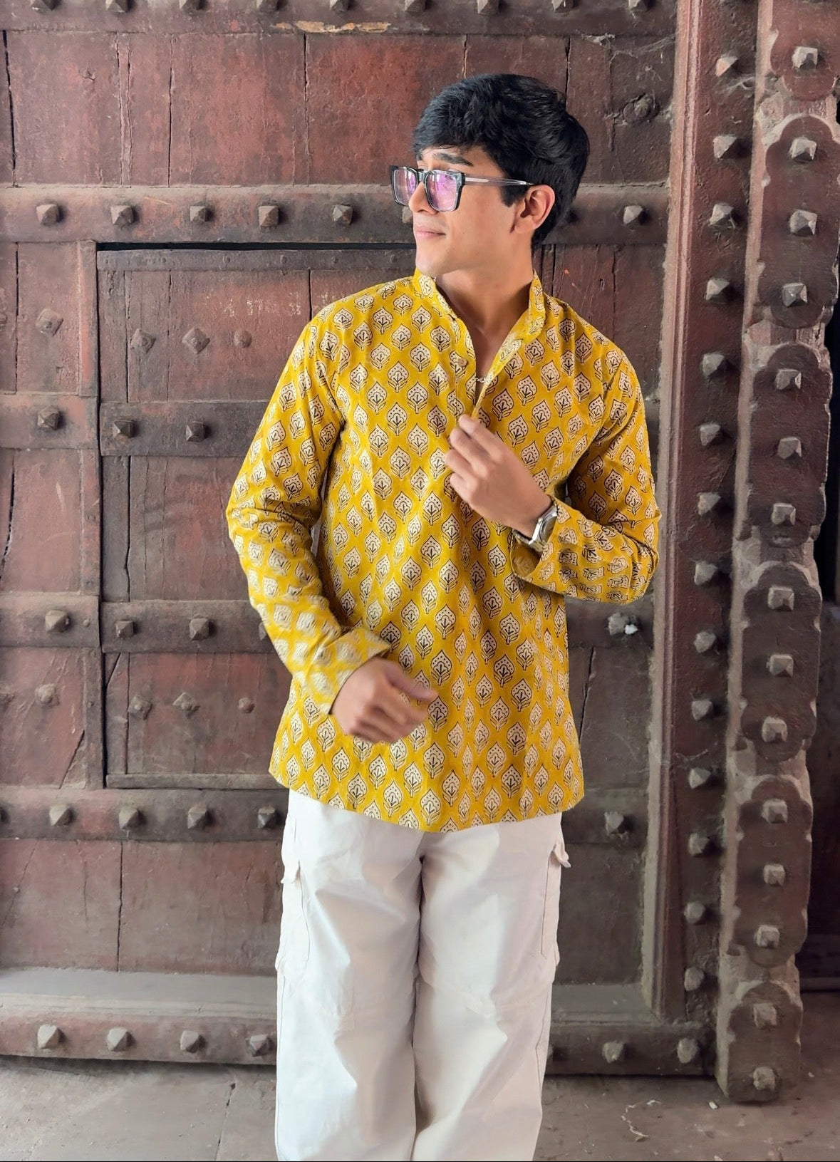 Person wearing a yellow patterned shirt and white pants standing in front of a large wooden door.