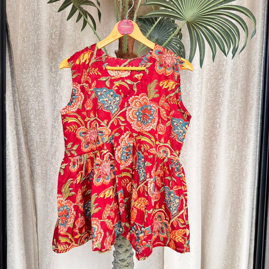 A scarlet red floral block print cut sleeves top displayed on a hanger with a tropical background.