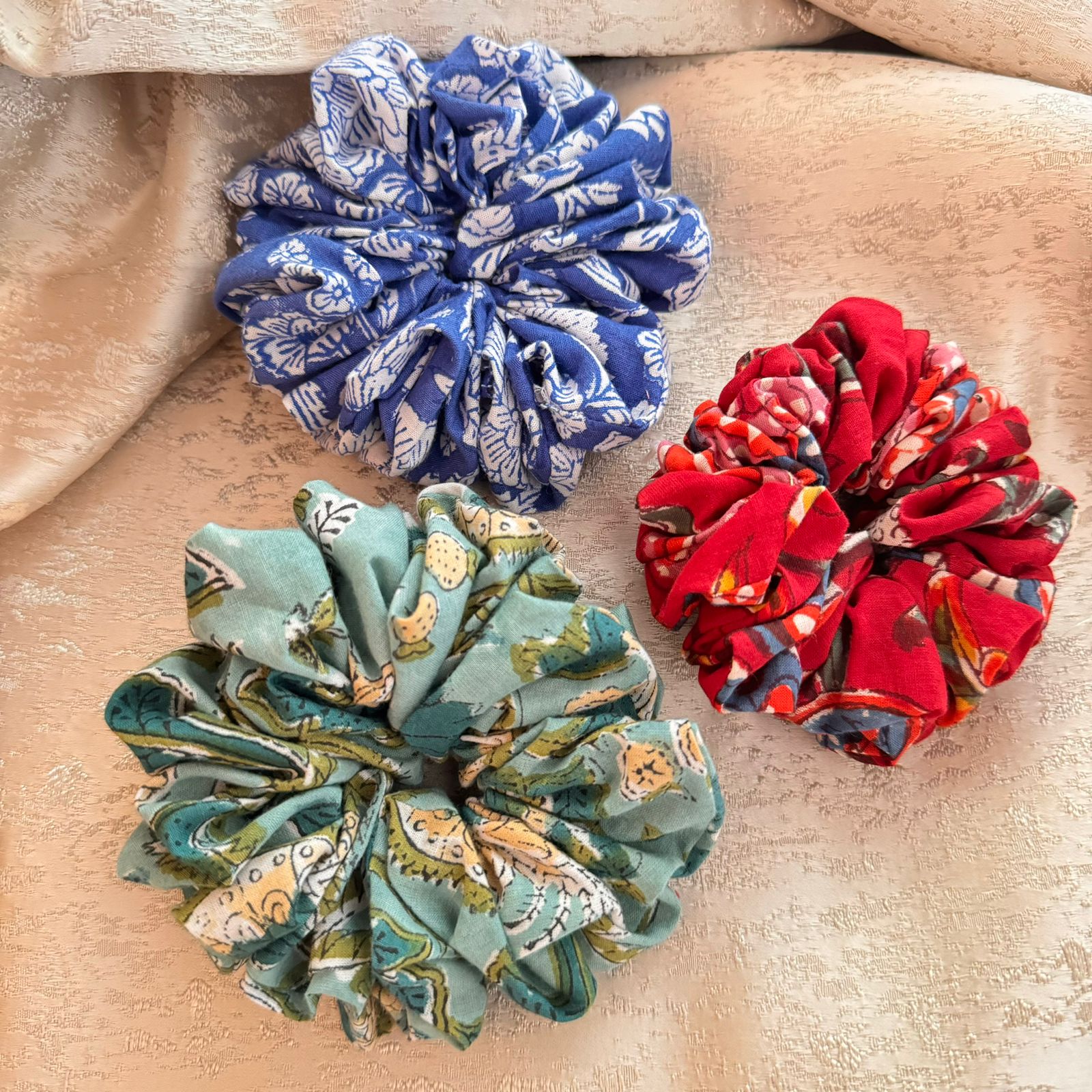 Three scrunchies on a textured background, one with a blue and white pattern, one with a red and black pattern, and one with a green and multicolor pattern.