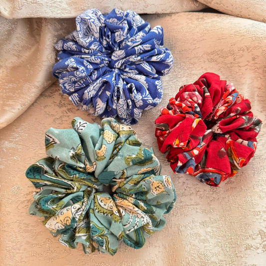 Three scrunchies on a textured background, one with a blue and white pattern, one with a red and black pattern, and one with a green and multicolor pattern.