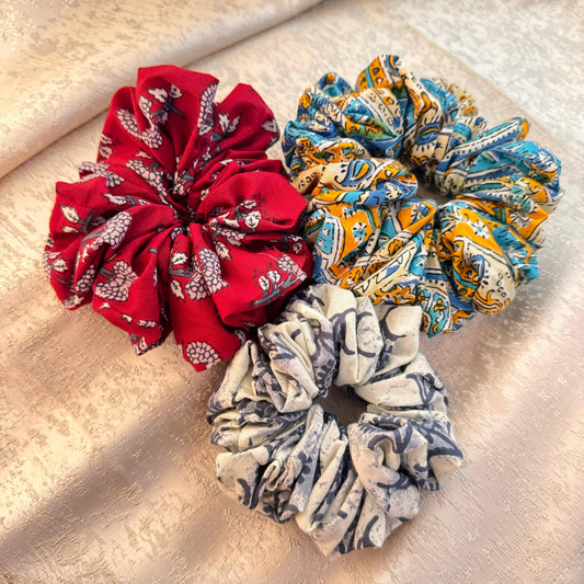 Three ponytail holders made of cotton, displayed on a textured fabric background. One scrunchy is red, the second is blue with multicolor patterns, and the third is gray with a floral design.