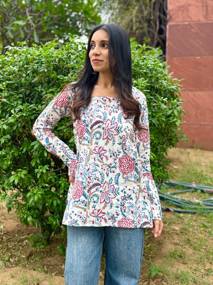 Woman wearing a floral blouse and jeans standing outdoors with greenery and a brick wall in the background.