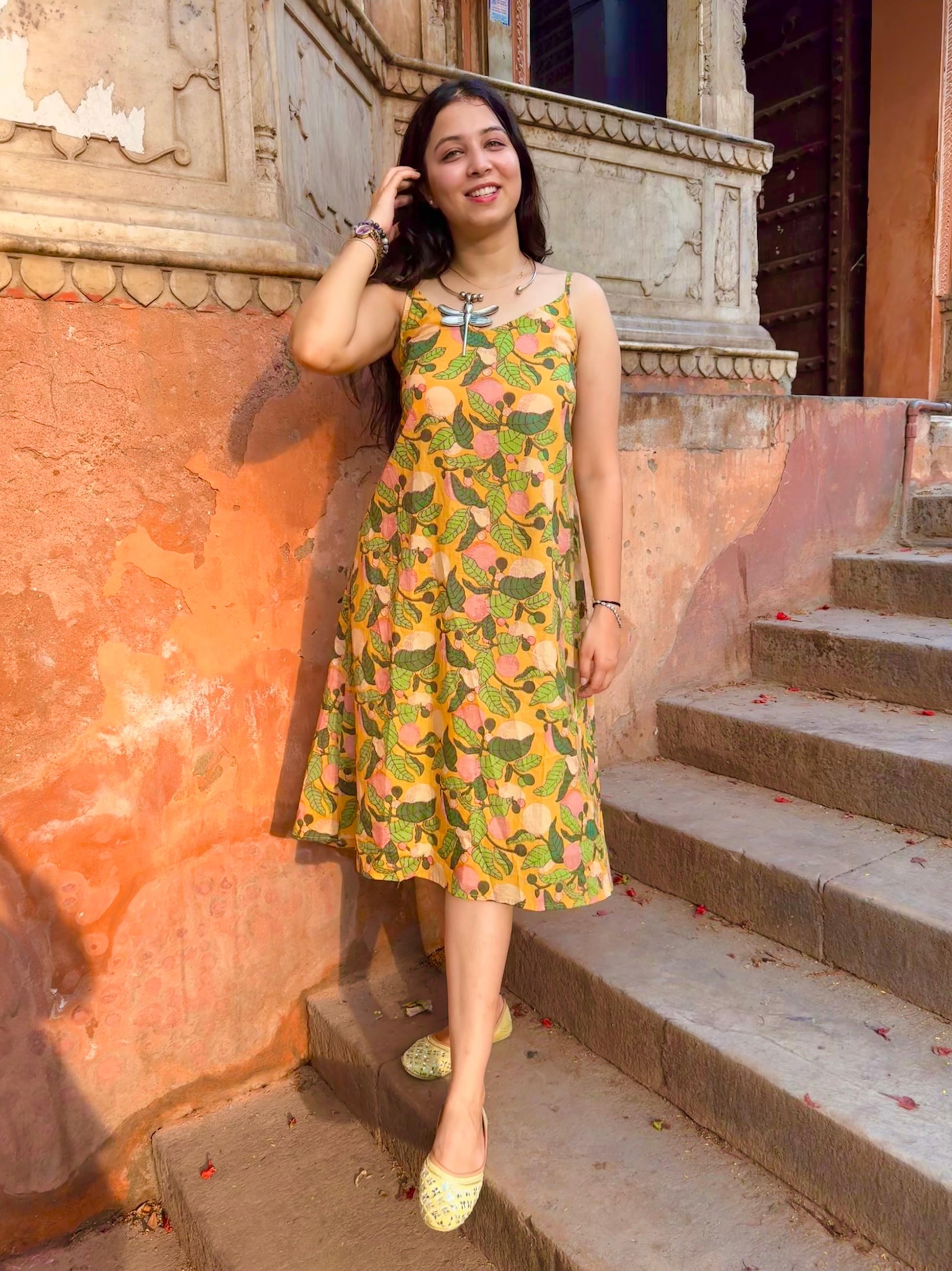 A woman standing on a set of stairs, wearing a vibrant yellow and green floral dress with 3/4 length sleeves and a V-neckline, posing with one hand on her hip.