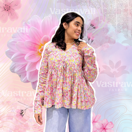 A woman standing with a smile on her face, wearing a floral pink top with long sleeves and a round neckline, paired with blue jeans. The background is adorned with pink and white flowers and the brand 'Vastravali' is visible.