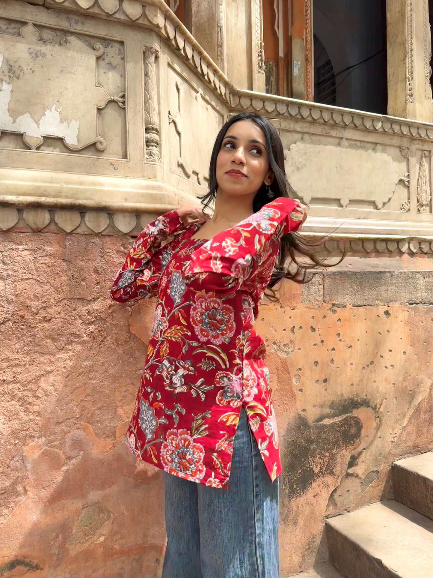 Woman in a red floral blouse standing against an ornate wall.
