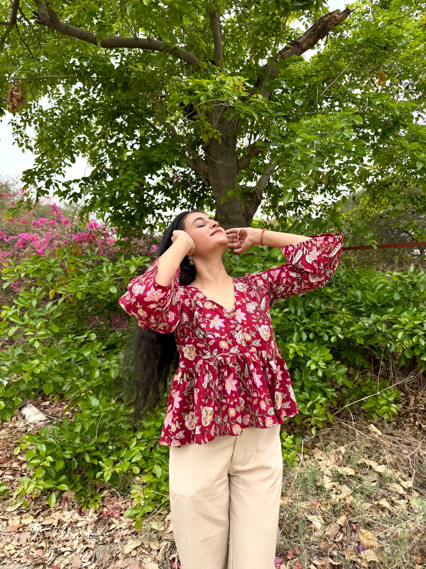 Woman in a red floral blouse standing outdoors with greenery and flowers in the background