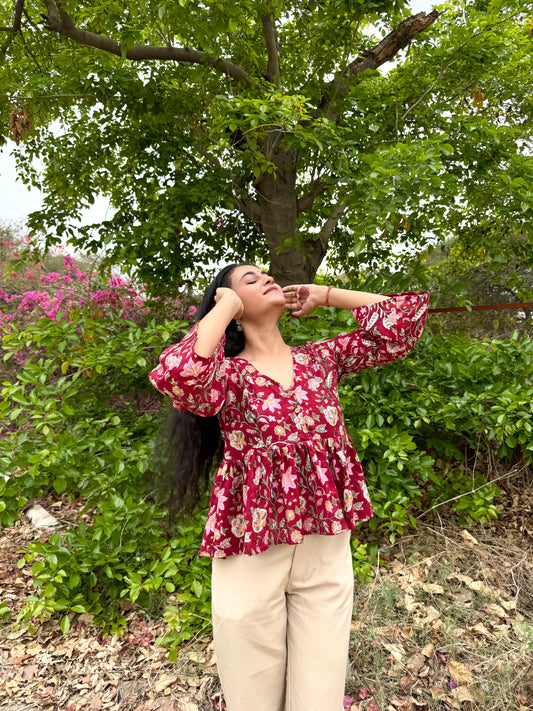 Woman in a red floral blouse standing outdoors with greenery and flowers in the background