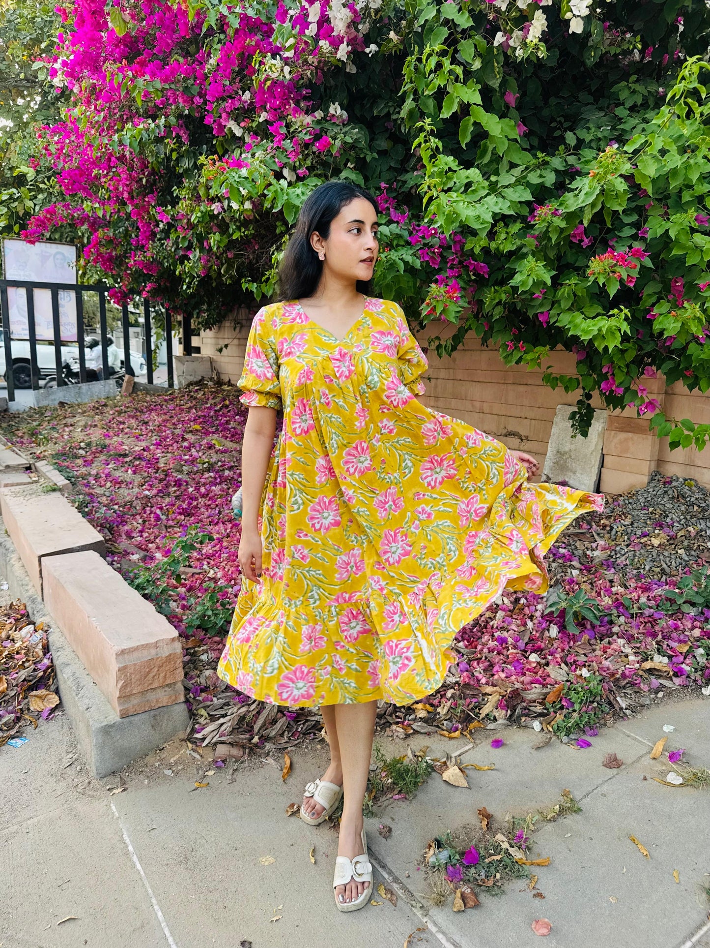 Woman in a yellow floral dress standing in front of flowering bushes.