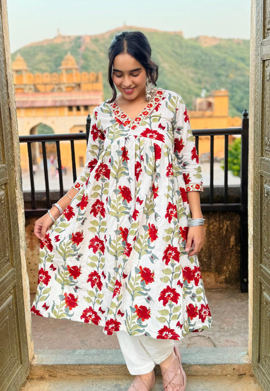 A woman standing on a balcony wearing a white, red, and green floral printed kurti with three-quarter sleeves and a maxi-length skirt.