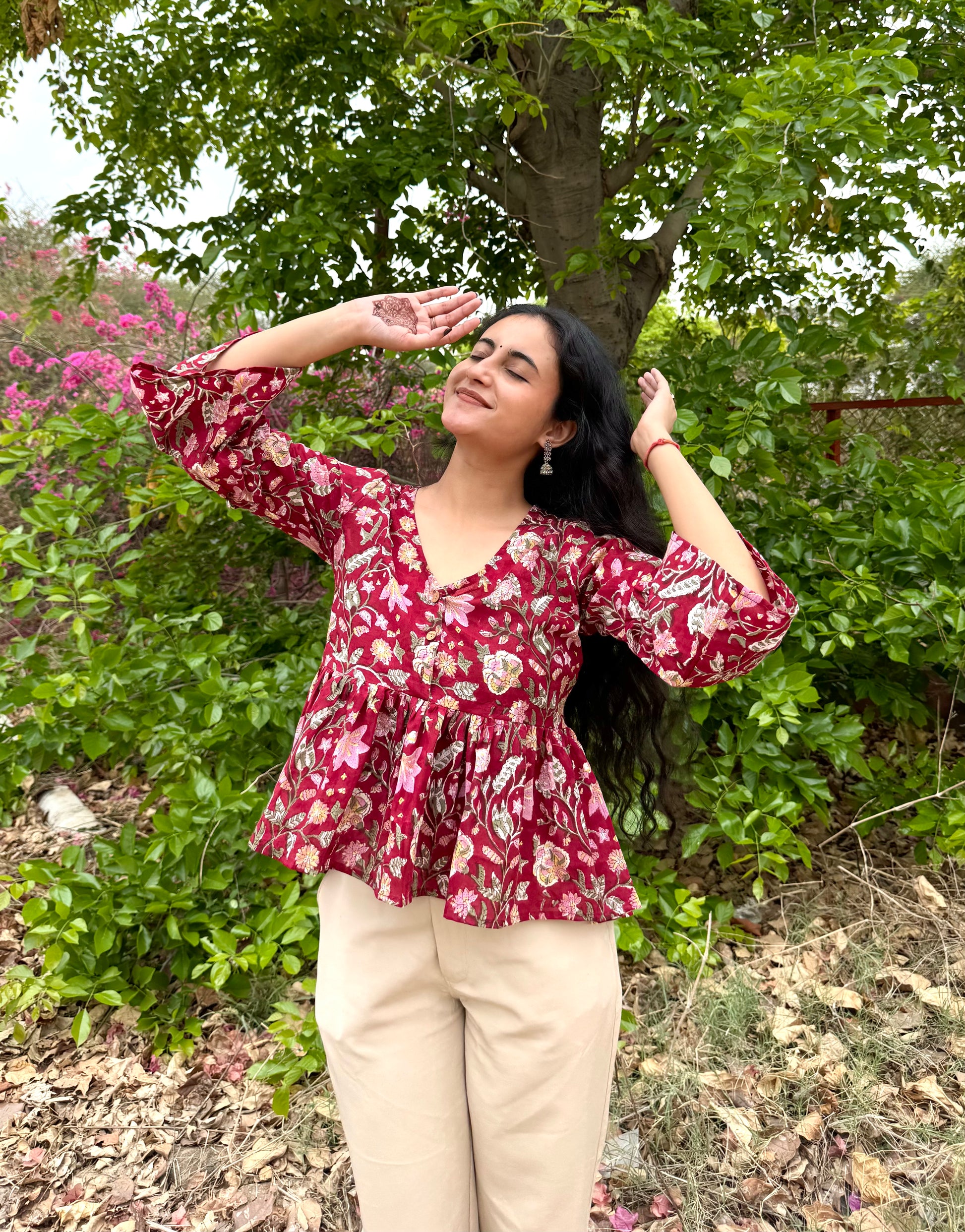 Woman in a red floral blouse standing outdoors with greenery in the background