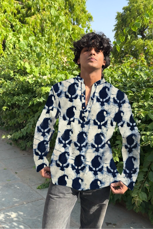 Person wearing a blue and white patterned shirt standing outdoors with greenery in the background