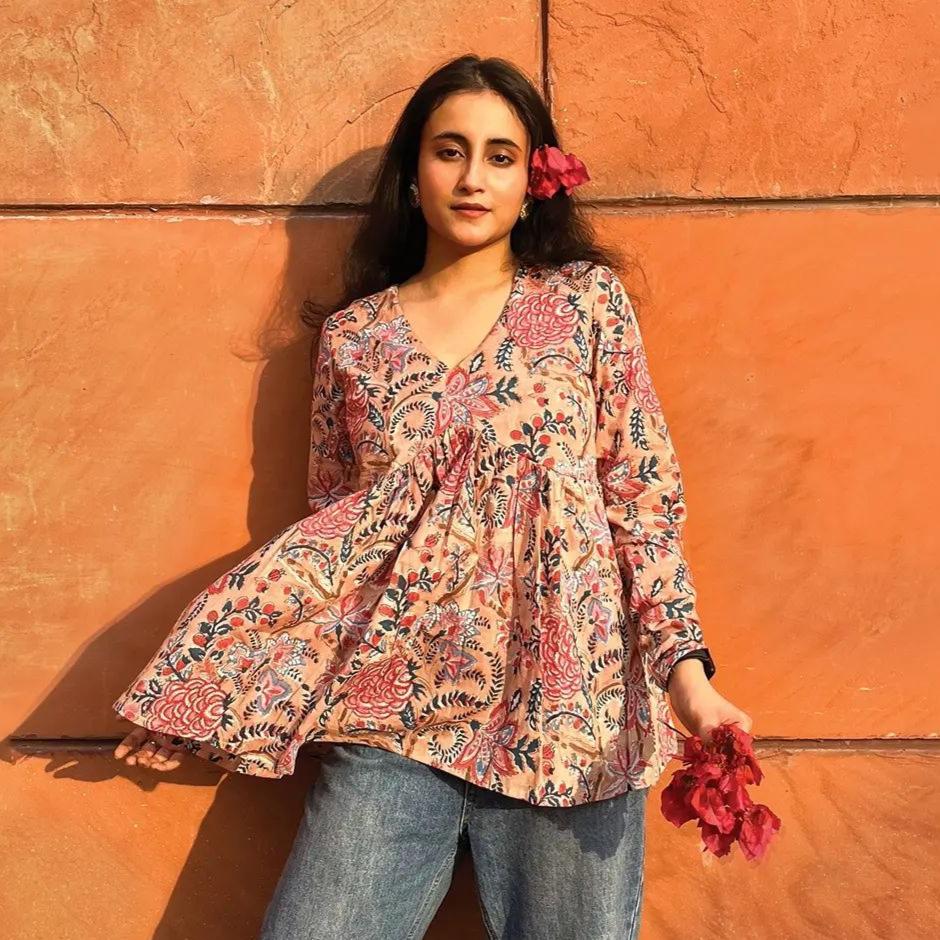 A woman wearing a beige cotton top with floral patterns, paired with blue jeans, standing against an orange wall.